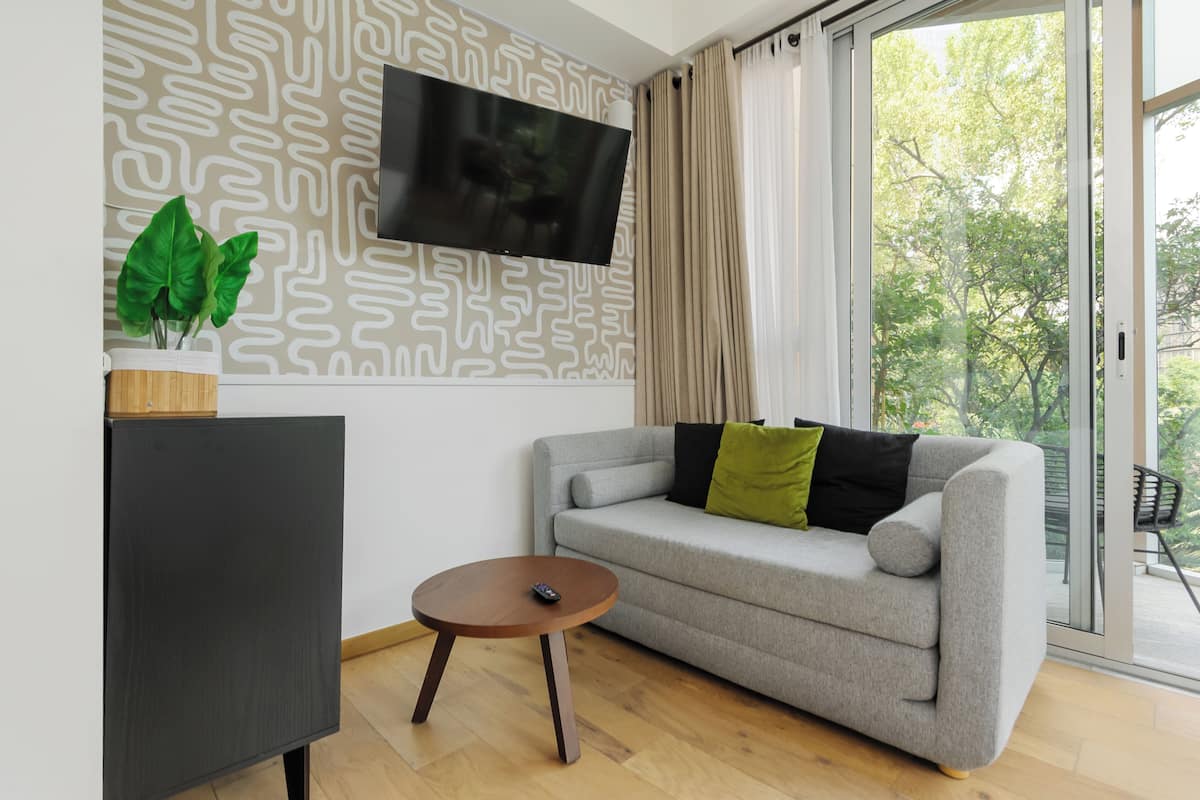 A modern living room with a grey sofa, a small round wooden table, a TV mounted on the wall, and a potted plant.