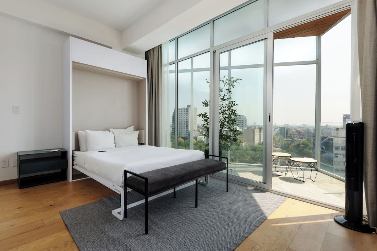 A modern bedroom with a large bed, a bench, a TV, and a balcony with a city view.
