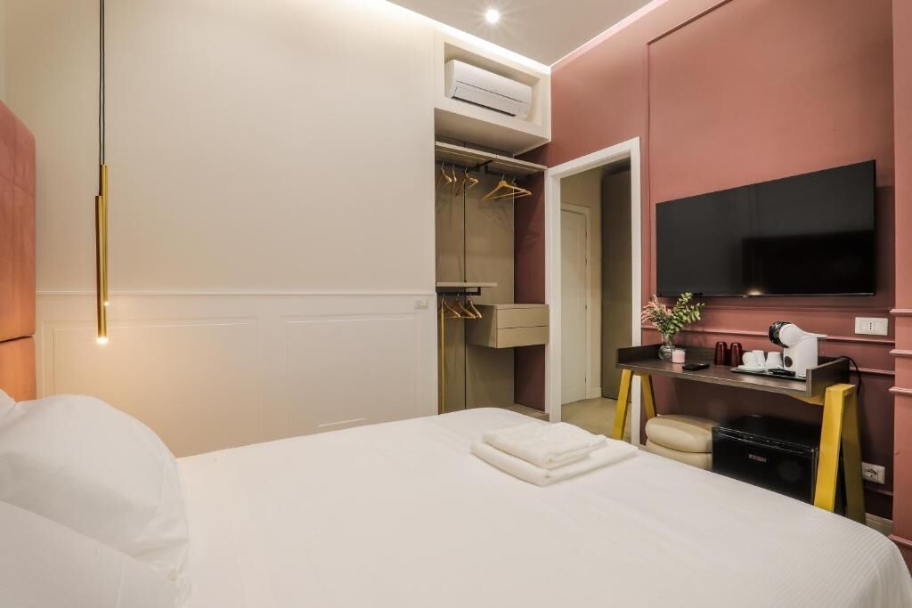 A hotel room with a bed, a TV mounted on the wall, a desk with a hairdryer, and a wardrobe with hanging clothes.