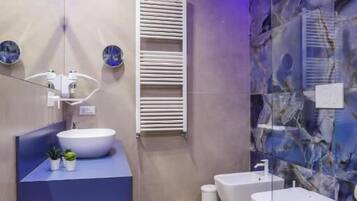 A modern bathroom with a large white sink, a blue vanity, a glass shower enclosure, and a marble wall.