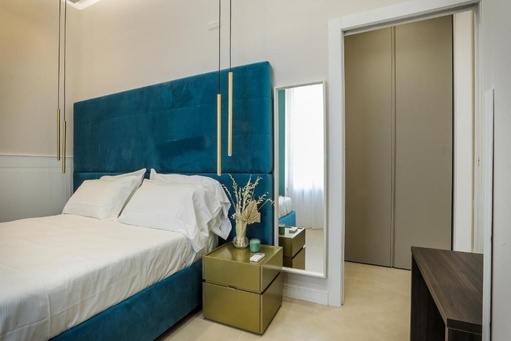 A modern bedroom with a blue headboard, a bed with white linens, a bedside table with a vase, and a mirror reflecting the room.