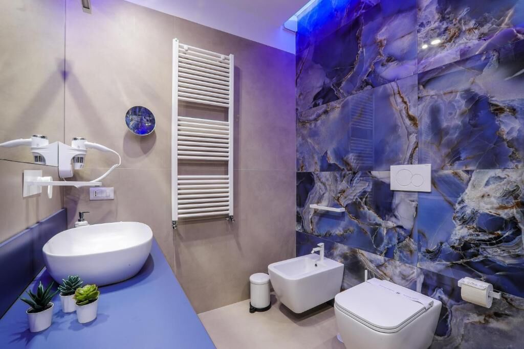 A modern bathroom with a large white sink, a bidet, and a toilet. There are small potted plants on the counter.