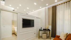 A modern living room with a flat-screen TV mounted on a white cabinet, a small round ottoman, a dark wooden side table with a lamp, and an orange sofa. - Conte Relais Suite (Naples)