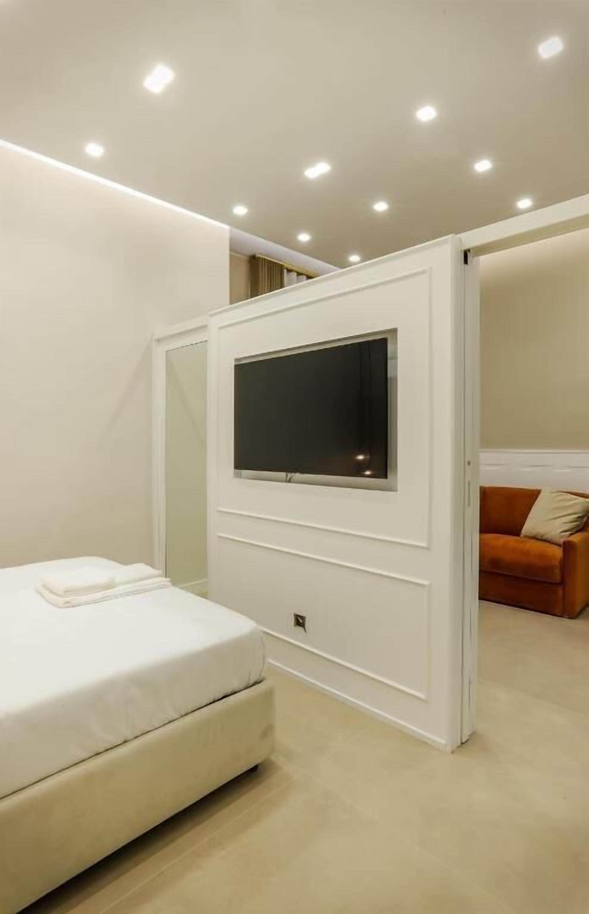 Una camera da letto moderna con un letto, una TV fissata a parete e una porta scorrevole che dà su un'altra stanza.