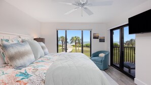 A bedroom with a large bed, a ceiling fan, a TV, and a view of the beach through the sliding glass doors.