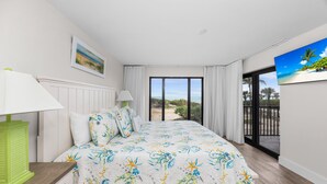 A bedroom with a large bed, a nightstand, a lamp, a painting, and a view of the outdoors through a sliding glass door.
