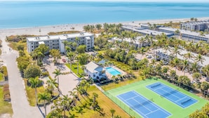 An aerial view of a coastal hotel with multiple buildings, a tennis court, and a beach in the background.
