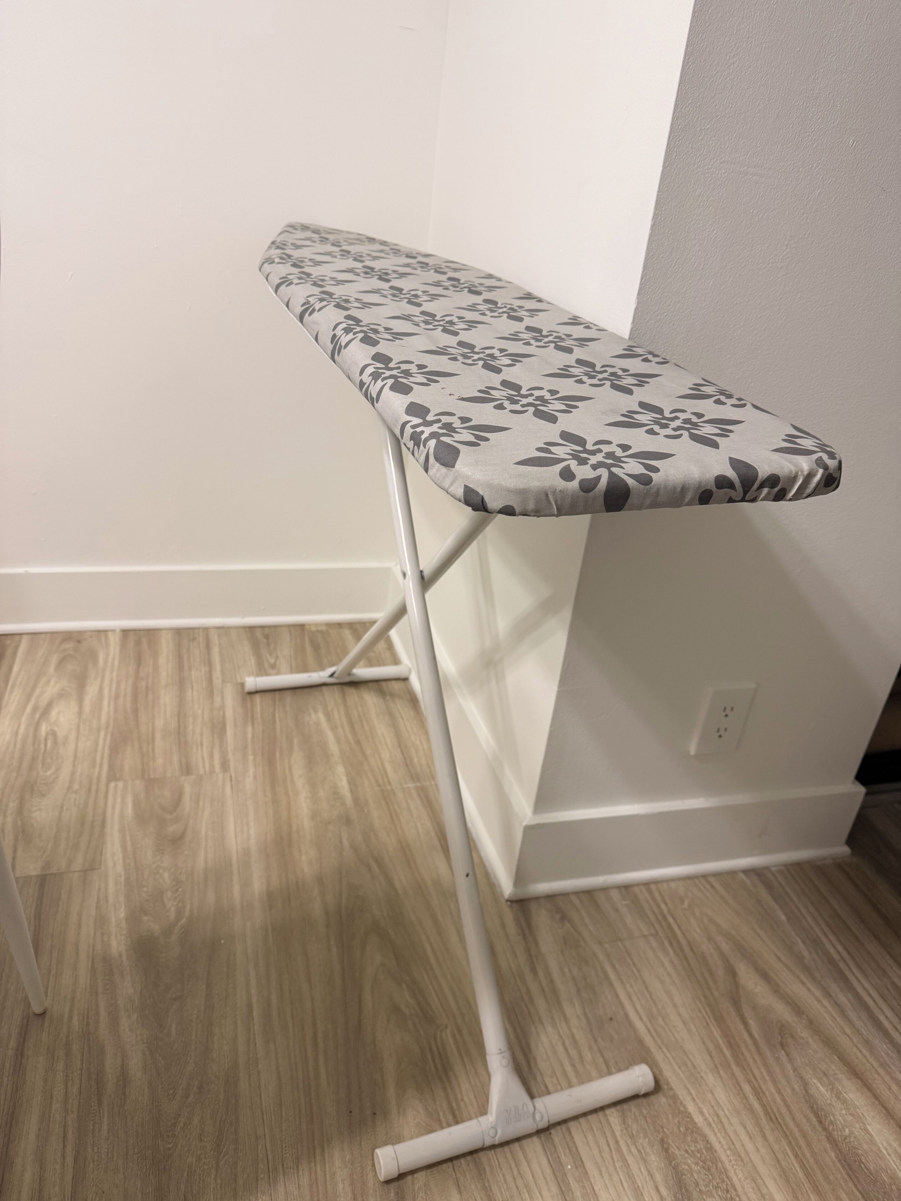 A corner ironing board with a floral pattern, positioned against a wall with a wooden floor.