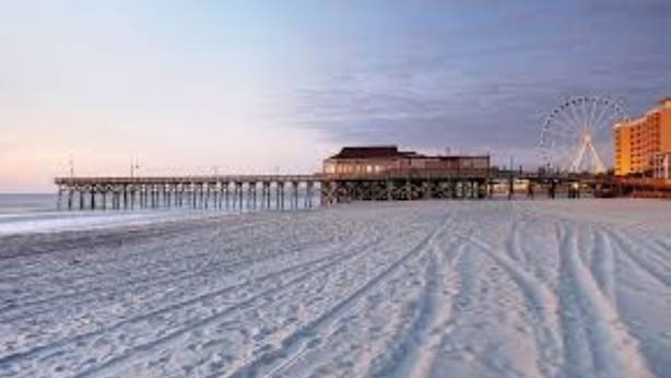 Een strand met een pier die het water in reikt, een groot gebouw en een reuzenrad op de achtergrond.