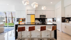 A modern kitchen with a central island, bar stools, white cabinets, and a large window overlooking a landscaped area. - Posh Private Panoramic View Compound w/Pool & Spa (Palm Springs)