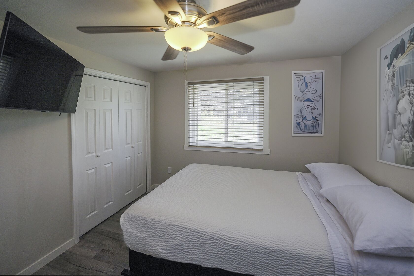 Une chambre à coucher comprenant un lit, un ventilateur de plafond, une fenêtre avec des stores, une porte et un téléviseur fixé au mur.