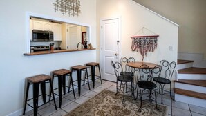 Een keuken met een magnetron, een spoelbak en een aanrecht. Er staan vier krukken bij een tafel met stoelen. Een decoratieve wandversiering is zichtbaar.