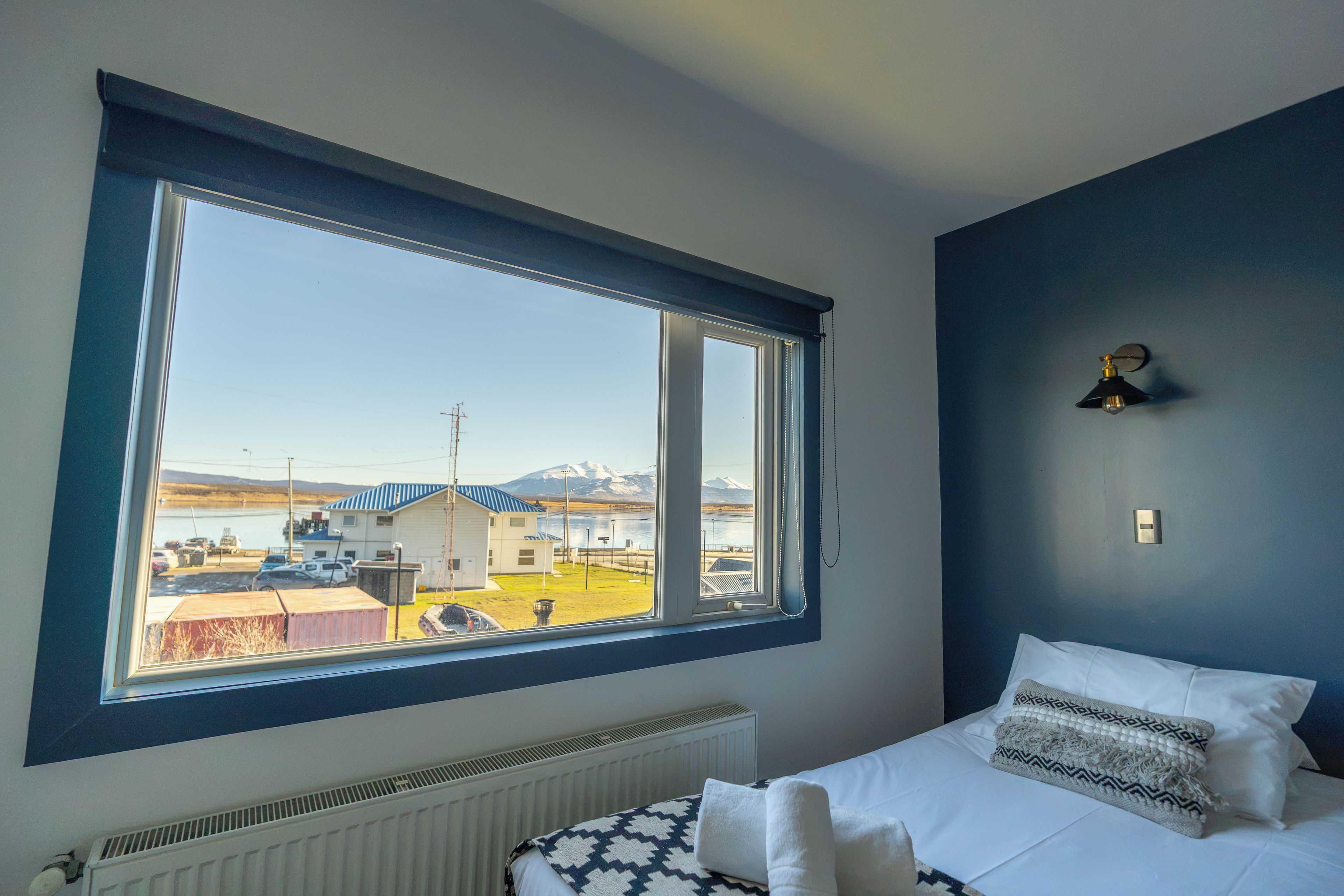 Ein Zimmer mit großem Fenster, einem Bett mit weißer Bettwäsche, einem Heizkörper und einer Wandleuchte.