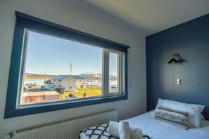 Een kamer met een groot raam, een bed met wit beddengoed, een radiator en een wandlamp.