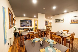 A dining area with wooden tables and chairs, a sofa, and a large window with a view. - Pristine Patagonia Hotel (Natales)