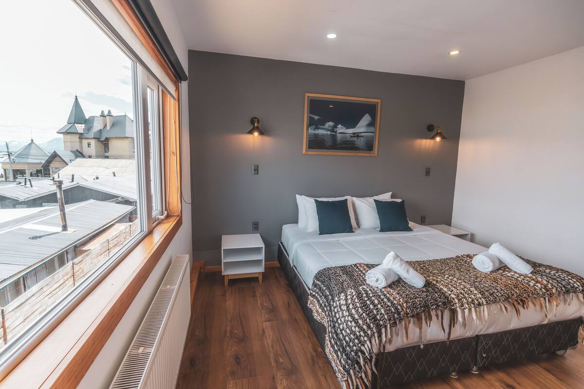 Ein Schlafzimmer mit großem Fenster, einem Bett mit gemustertem Überwurf, zwei Wandleuchten und einem gerahmten Bild an der Wand.
