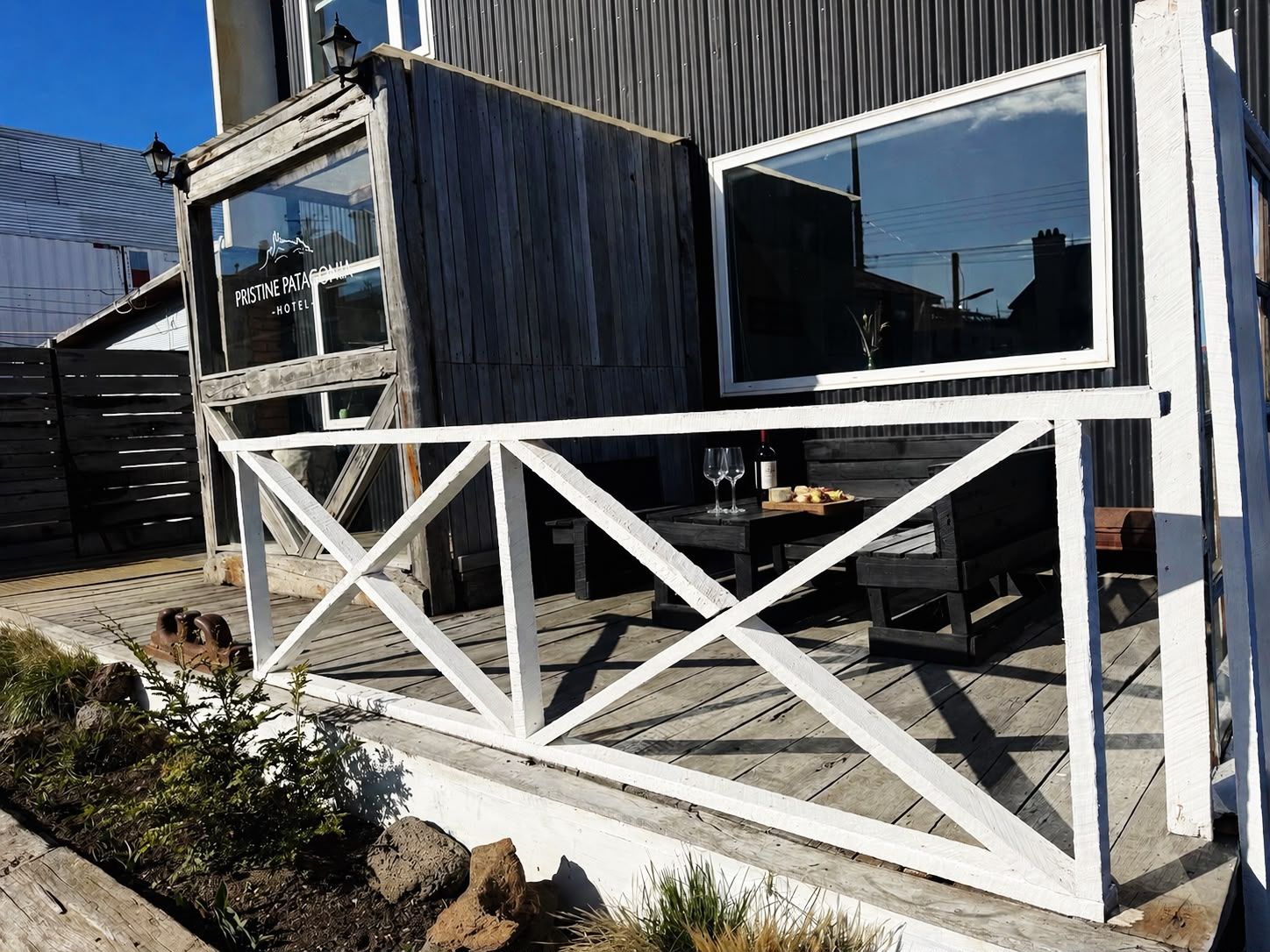 A wooden deck with a white railing, a table with a bottle and glasses, and a building with large windows.