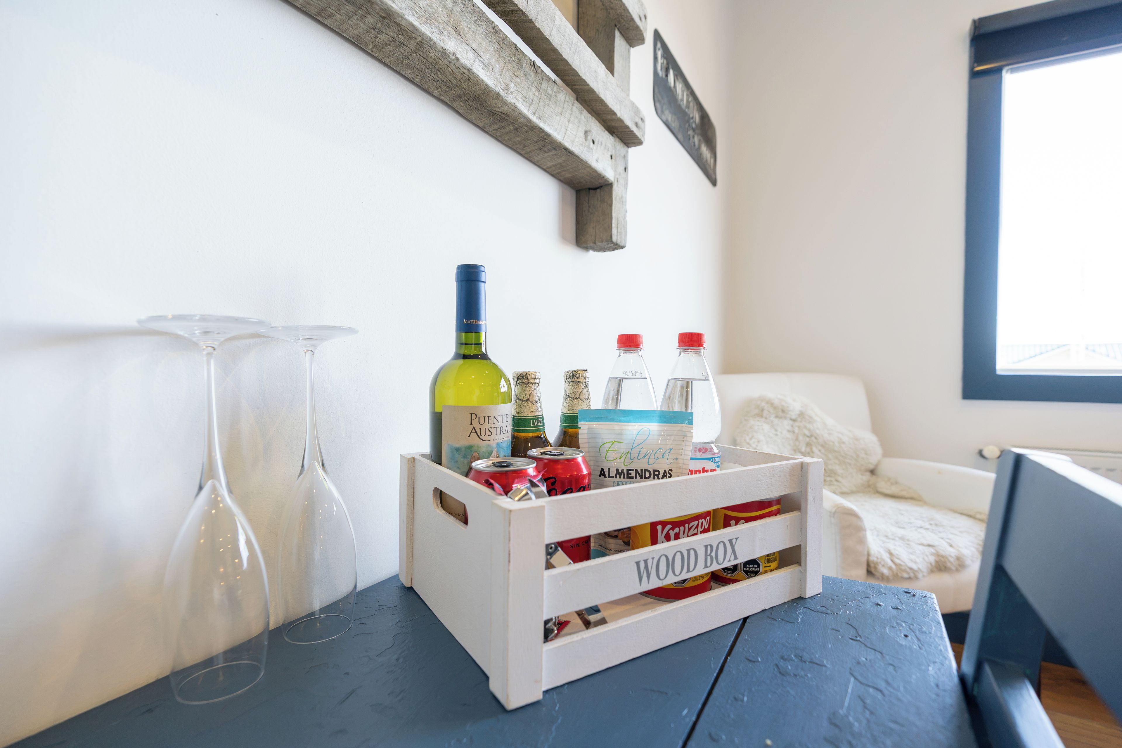 Una caja de madera con botellas y latas sobre una mesa, vasos en la pared y una ventana al fondo.