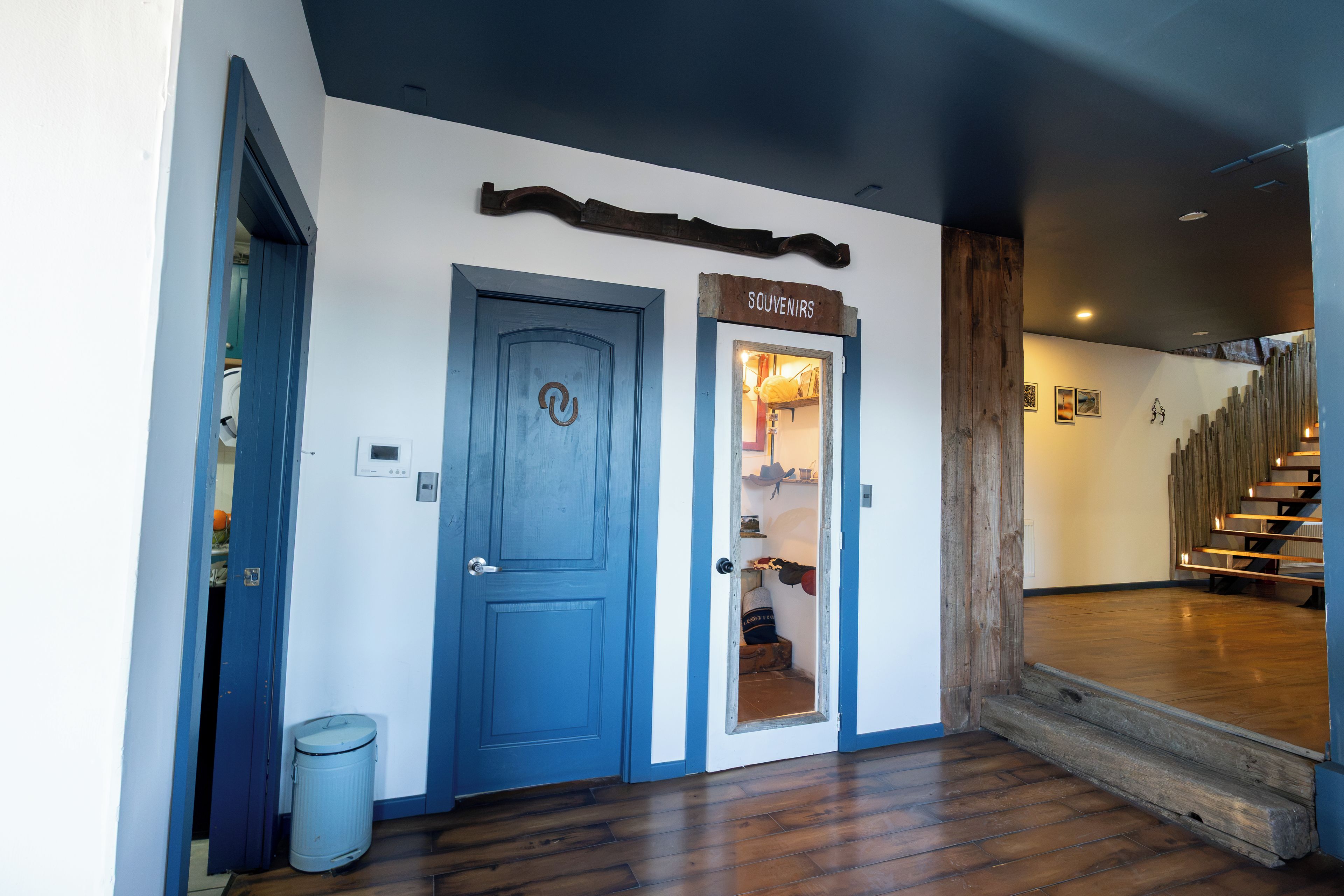 Un pasillo con una puerta azul que indica 'SOUVENIRS', un basurero y una escalera de madera.