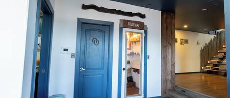 Un pasillo con una puerta azul que indica 'SOUVENIRS', un basurero y una escalera de madera.