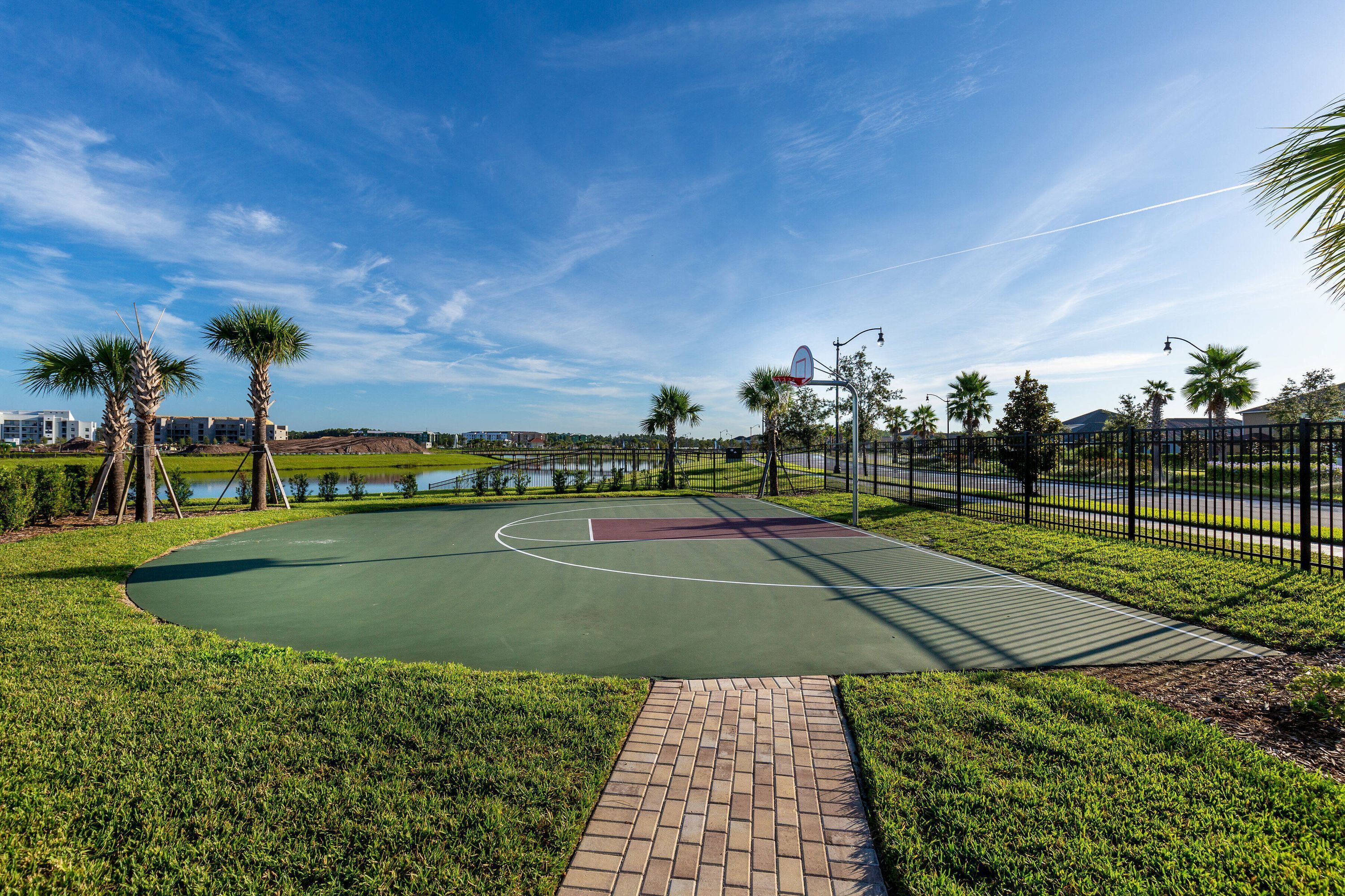 Una cancha de baloncesto con un camino de ladrillo que la conecta, rodeada de césped y palmeras.