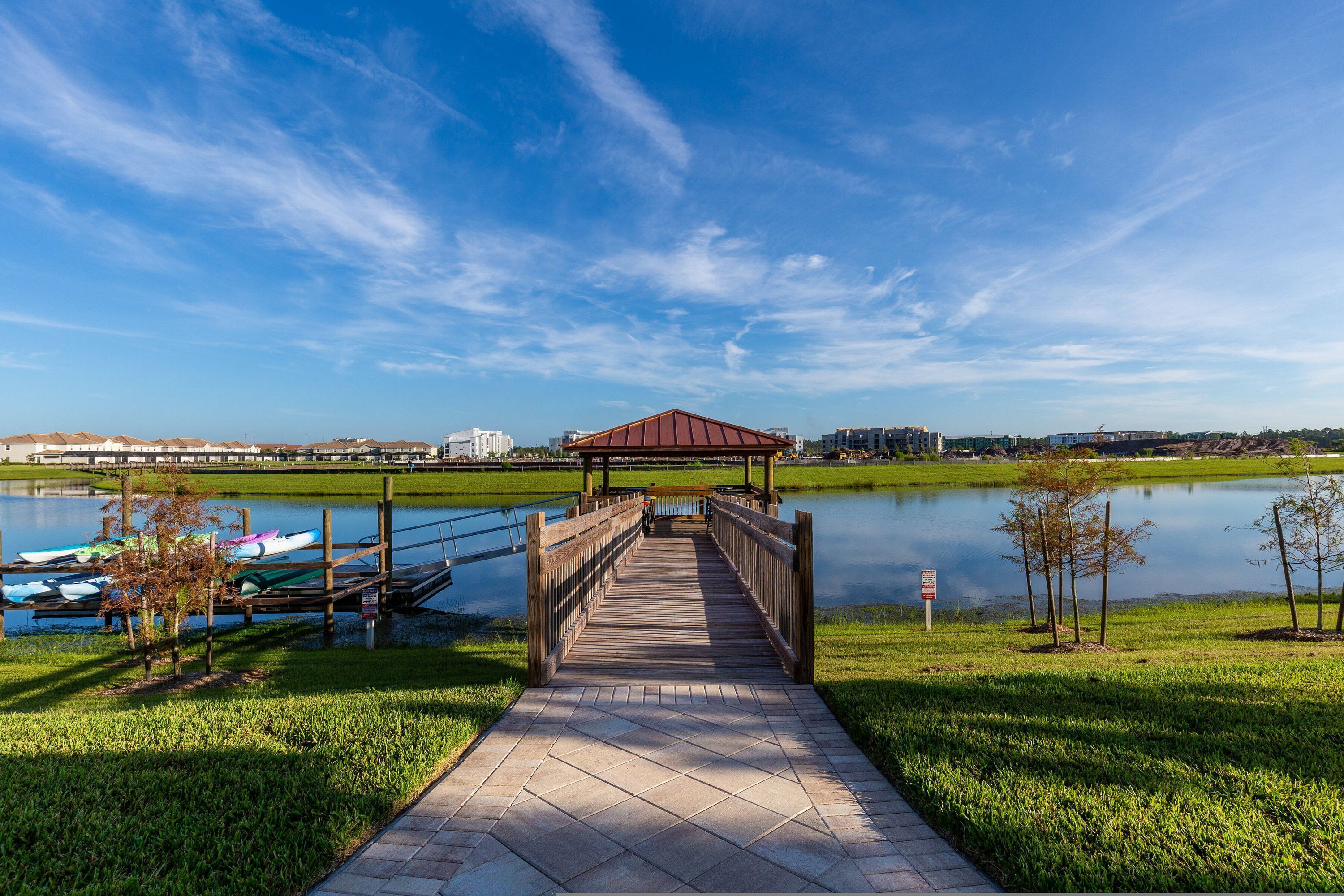 A wooden boardwalk leading to a gazebo over a calm lake, surrounded by grassy areas and boats.