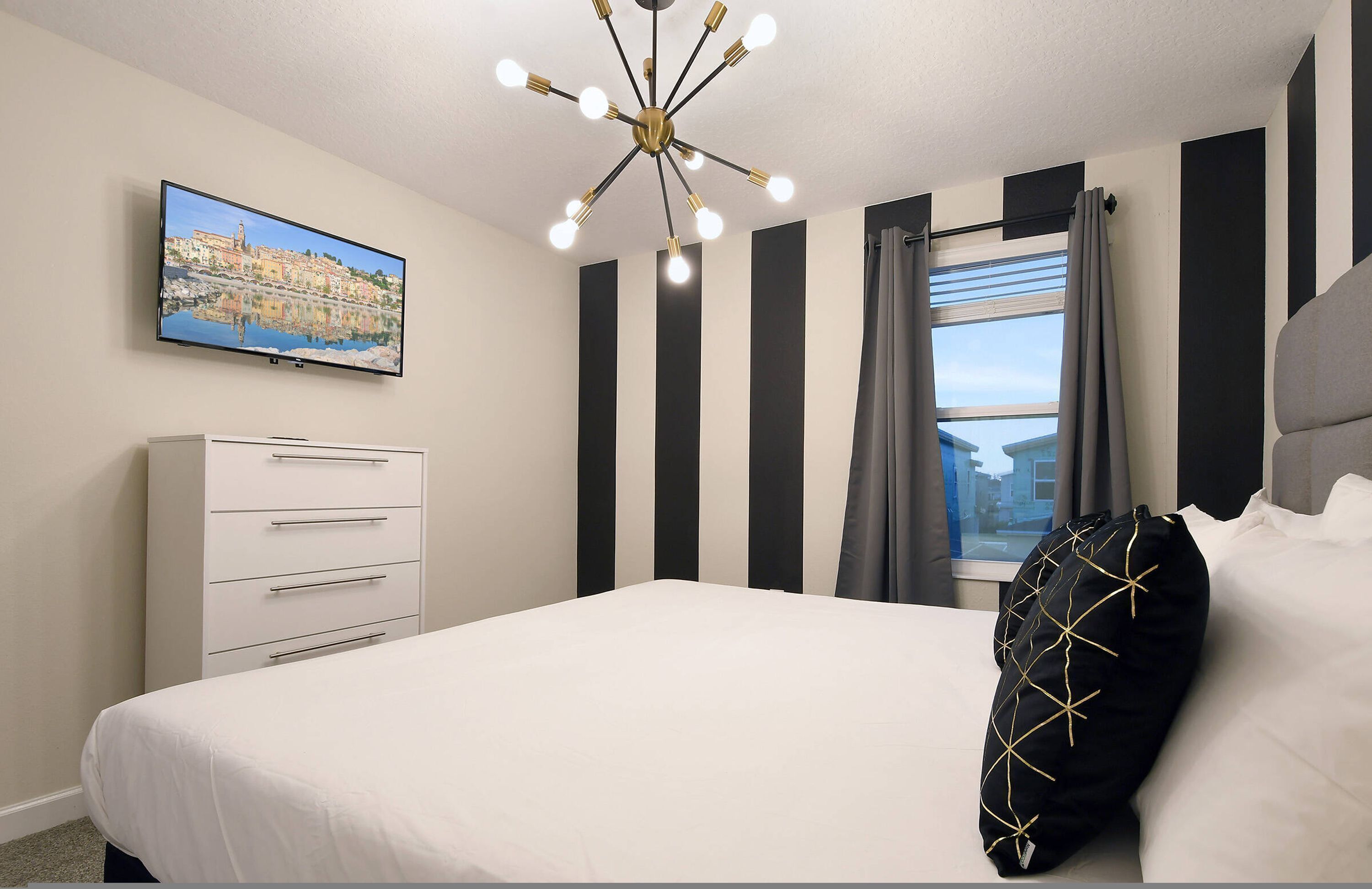 A bedroom with a bed, a dresser, a TV mounted on the wall, and a window with curtains.