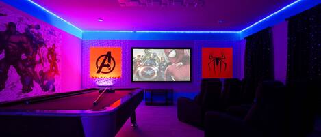 A room with a pool table, a large screen, and wall art featuring superhero characters.