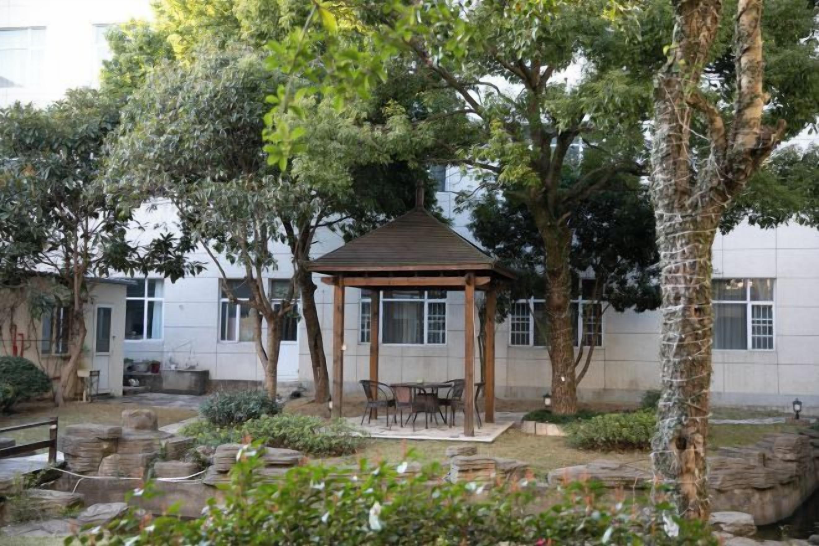 A gazebo with a roof and four chairs, surrounded by trees and a small water feature.