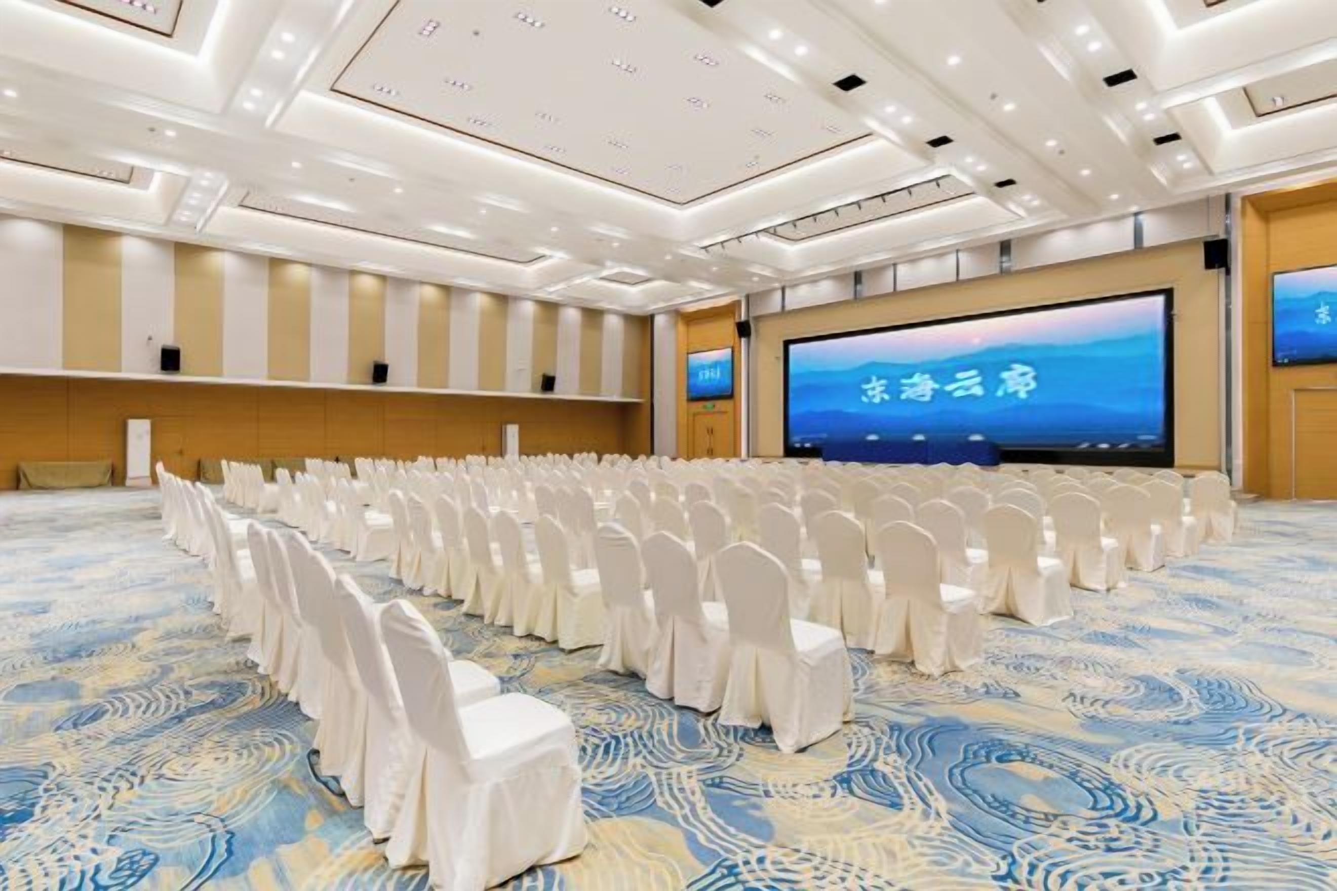 Un salón de conferencias con filas de sillas blancas, una pantalla grande y una alfombra estampada.