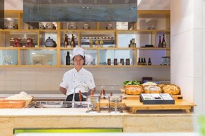 Un chef dans une cuisine, avec un comptoir où sont exposés du pain et des pâtisseries, des étagères remplies de bouteilles et un mur recouvert de carreaux.