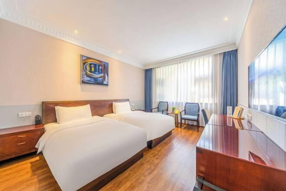 A hotel room with two beds, a desk, a chair, a painting, and a large window with curtains.