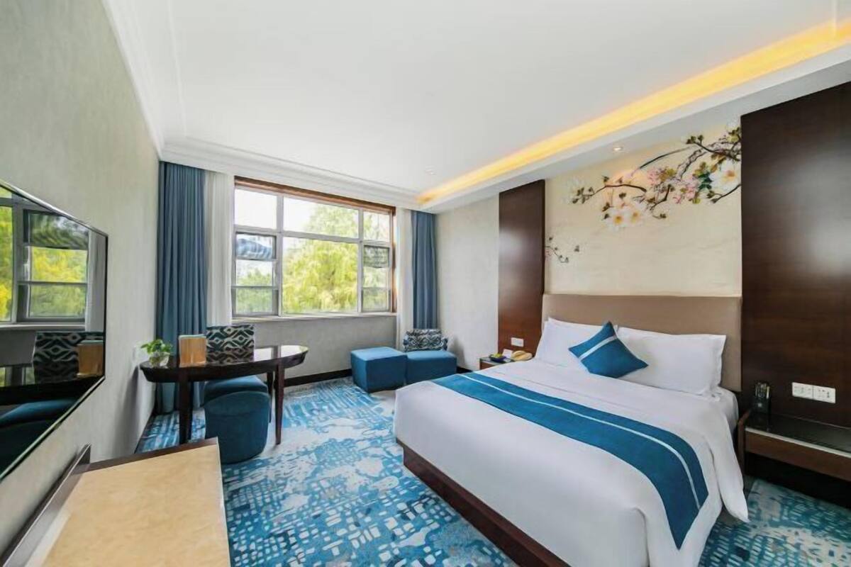 A hotel room with a large bed, a desk, a chair, a TV, and a window with a view of trees.