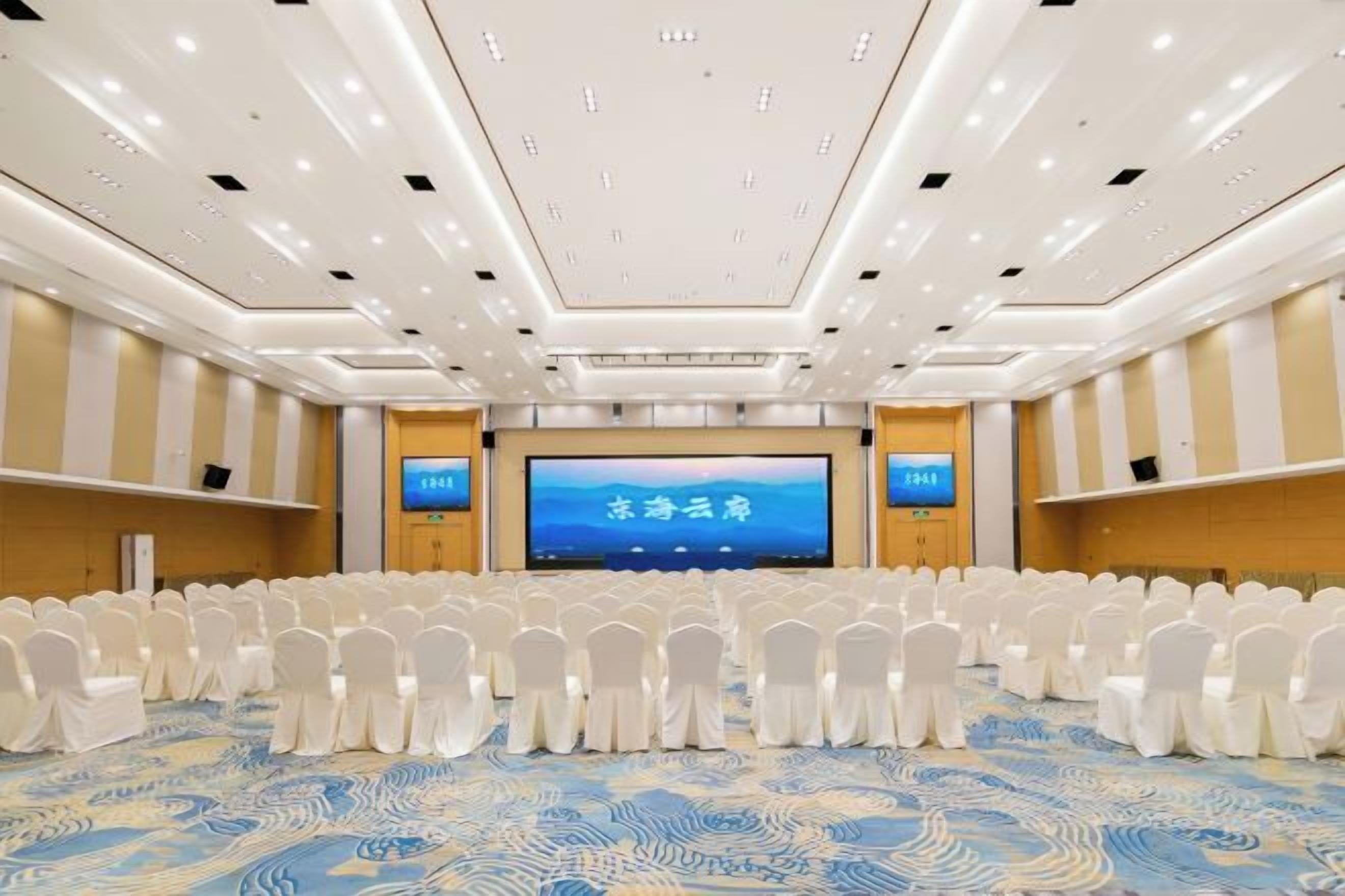 A conference room with rows of white chairs, a large screen at the front, and a patterned carpet.