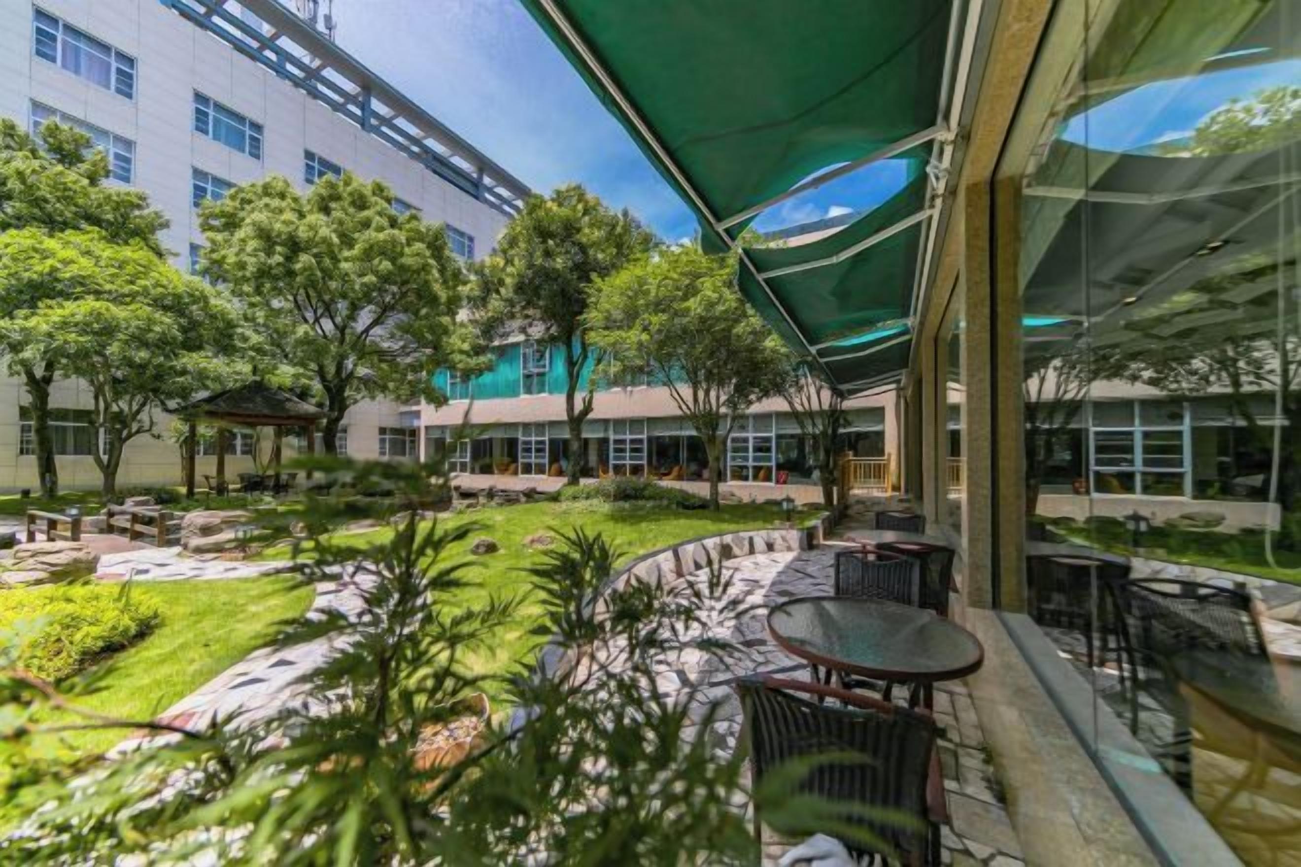 An outdoor seating area with tables and chairs, surrounded by greenery and a building in the background.