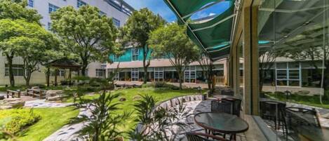 An outdoor seating area with tables and chairs, surrounded by greenery and a building in the background.