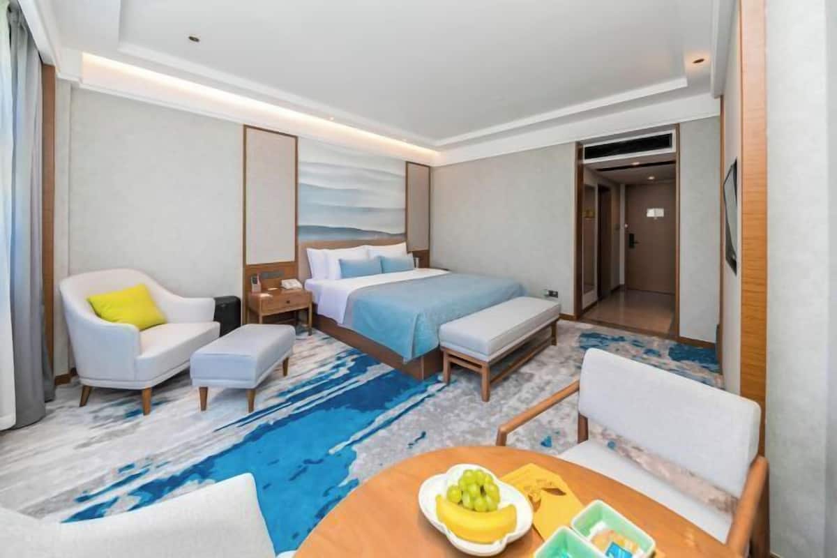 A hotel room with a bed, a desk, a chair, a table with fruit, and a large window.