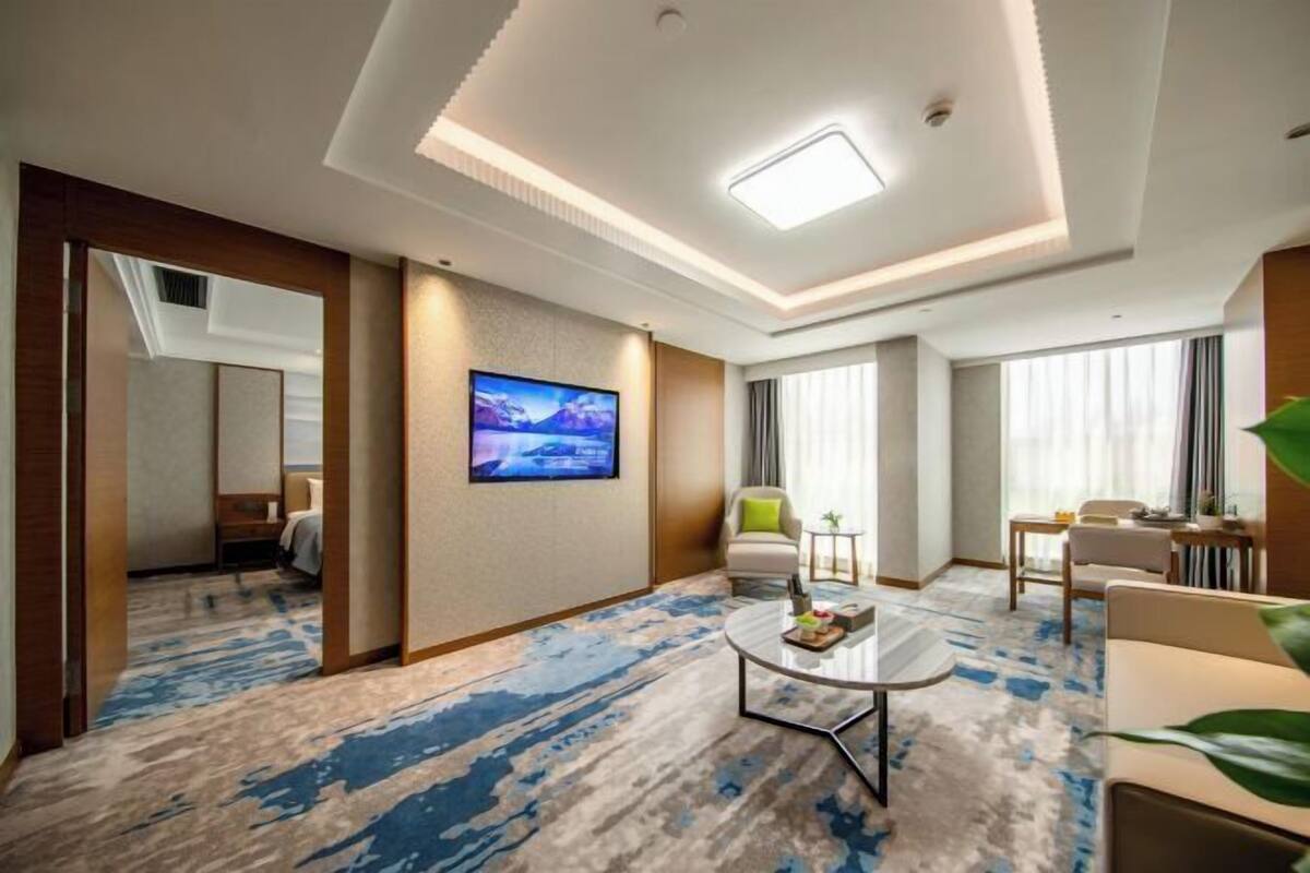 A hotel room with a large flat-screen TV, a round coffee table, a sofa, a chair, and a desk with a lamp.