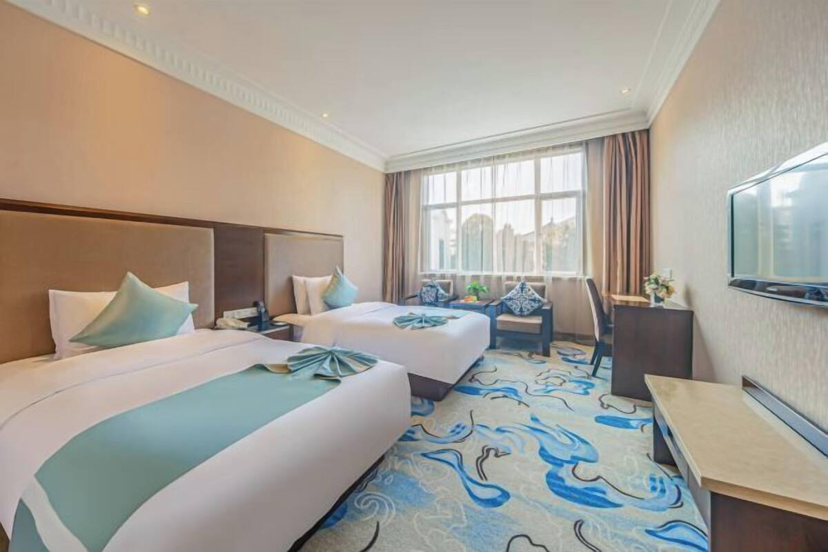 A hotel room with two beds, a desk, a TV, and a window with curtains.