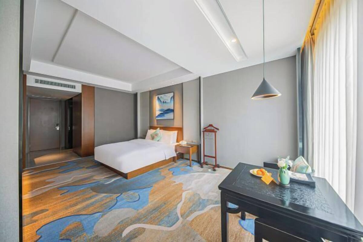 A hotel room with a bed, bedside table, lamp, and a view of the outdoors.
