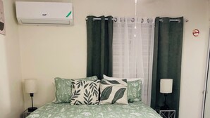 Un dormitorio con colcha verde estampada, almohada blanca, lámpara de noche y ventilador de techo.