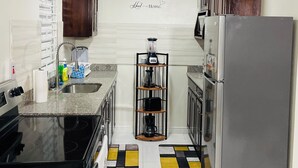 Una cocina con estufa negra, refrigerador de acero inoxidable y microondas empotrado. Se observa un aparador con utensilios de cocina y un tapete con diseño en el suelo.