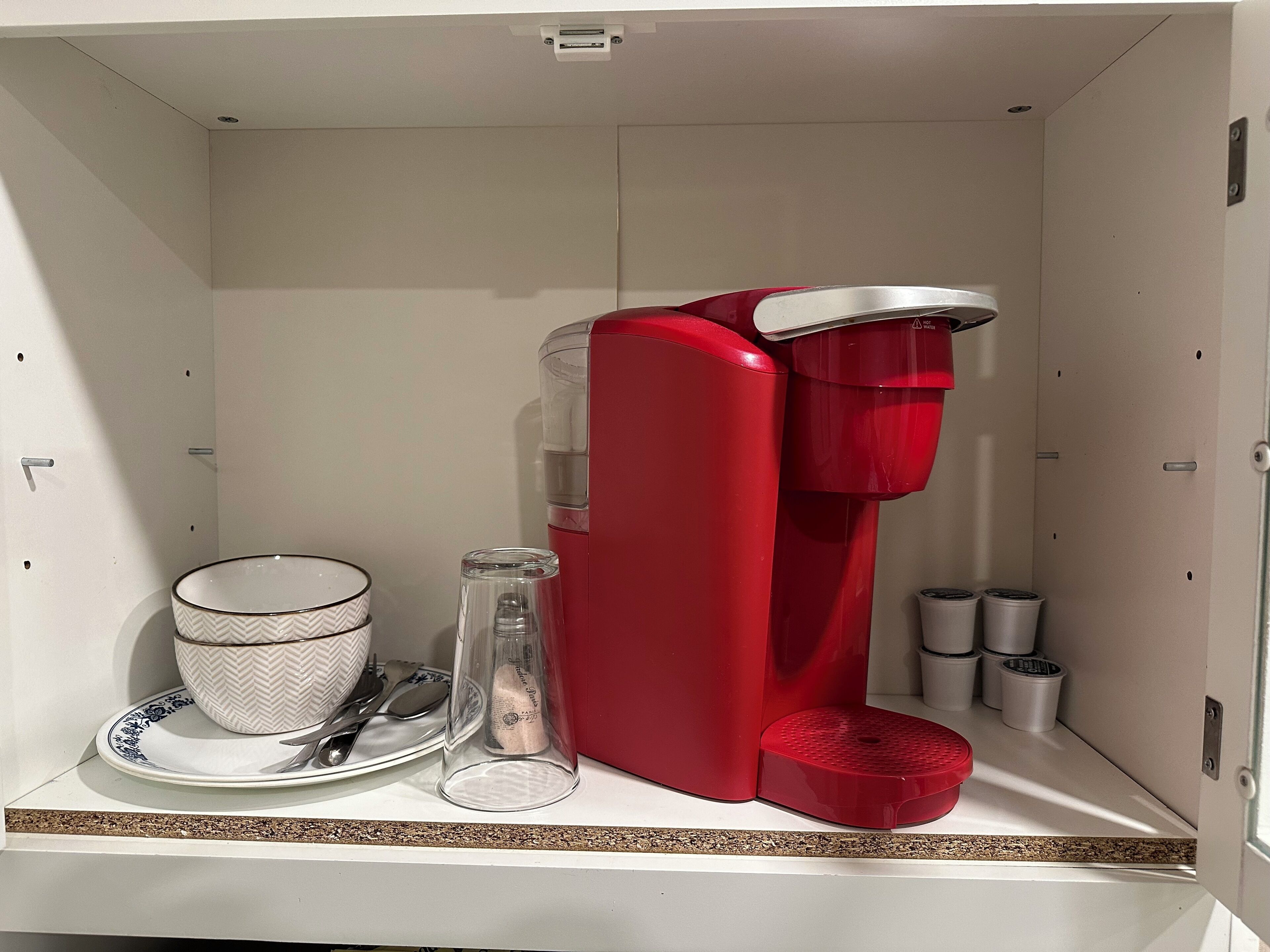 Et lille tekøkken med en rød kaffemaskine, en vandflaske i glas, en hvid kop og en tallerken med bestik.