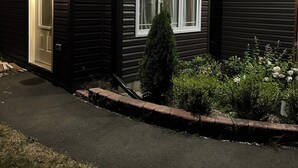 A two-story house with a dark exterior, a lit entrance, and a small garden with various plants and flowers. - Charming small studio in adorable St. John's WiFi access, 2 twin beds, full bath (St. John's)