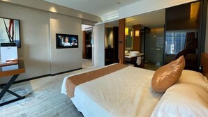 Hotellihuone, jossa on sänky, televisio, työpöytä, peili ja kylpyhuone, joka näkyy avoimen oven läpi.