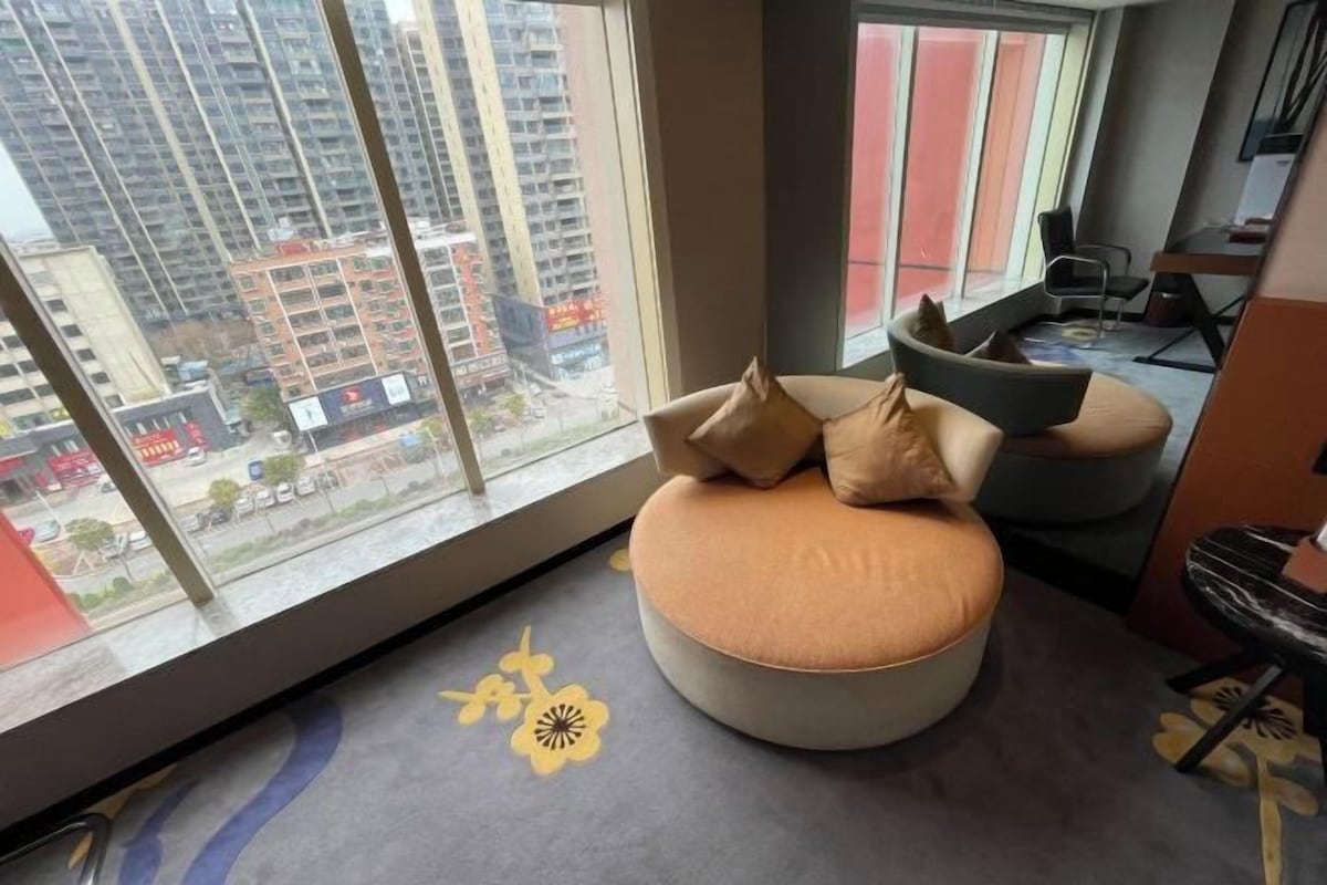 A modern room with a large window, a round seating area with cushions, and a desk with a chair.