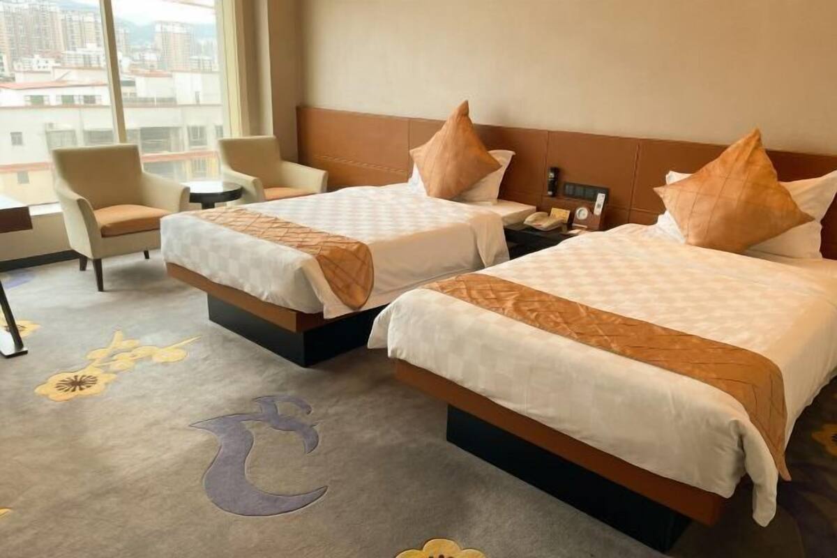 A hotel room with two beds, a sofa, a small table, and a view of buildings through large windows.