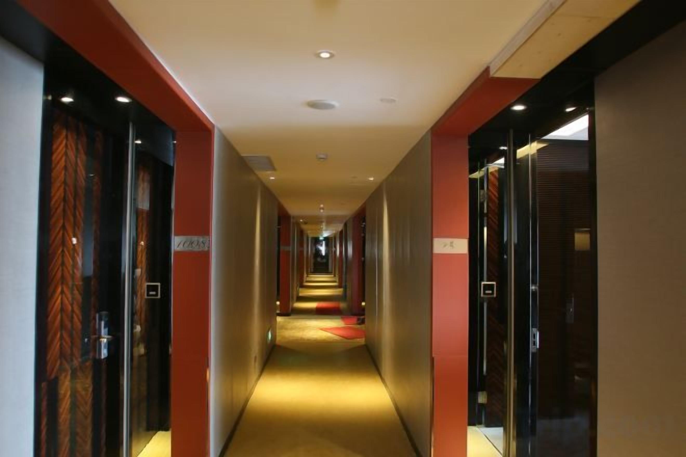Un couloir d’hôtel avec des portes des deux côtés, un sol recouvert de moquette et un tapis rouge menant au bout du couloir.