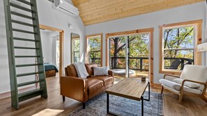 Un salon spacieux avec un plafond en bois, de grandes fenêtres donnant sur la verdure, un canapé en cuir marron, un fauteuil blanc et une table basse en bois.