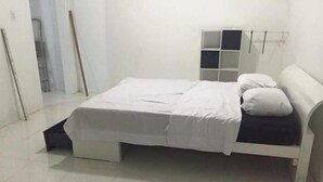 Een bed met een wit matras en kussens, een nachtkastje en een klein wandrek.